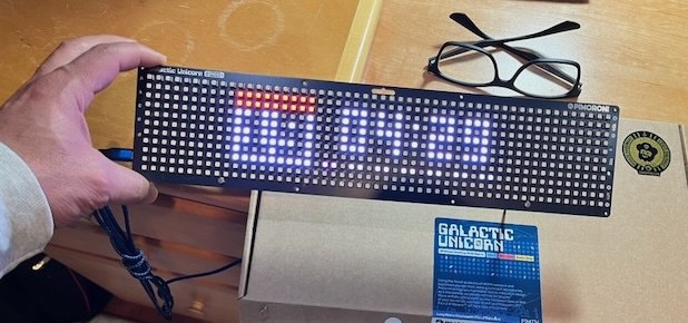 A hand holding the Galactic Unicorn: a long, thin black LED panel showing the time in white digits with a small red icon at the left end